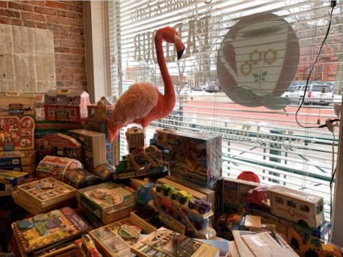 A pink flamingo plush toy stands among various vintage toys and games in a shop, with a window view outside.