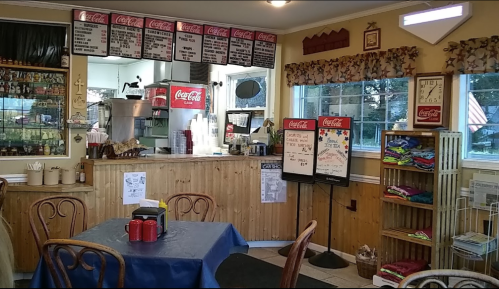 Cozy diner interior with a wooden counter, menu boards, colorful shirts for sale, and a blue tablecloth.