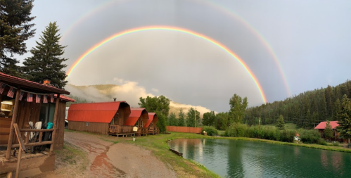 A vibrant double rainbow arcs over a serene pond and rustic cabins surrounded by lush trees and mountains.
