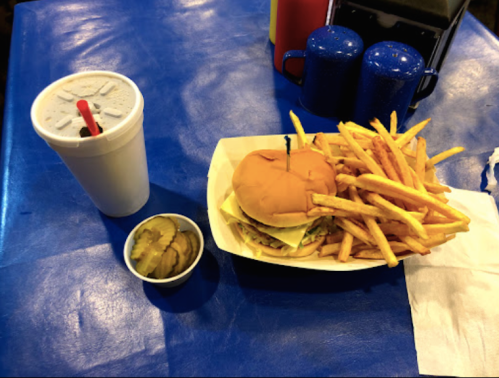 A burger with lettuce and tomato, a side of fries, a cup of soda, and a small container of pickles on a blue table.