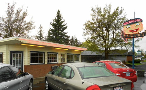 A retro-style burger restaurant with a neon sign, surrounded by parked cars and trees on a rainy day.