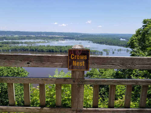 A wooden railing with a "Crows Nest" sign overlooks a scenic river and lush green landscape under a clear blue sky.