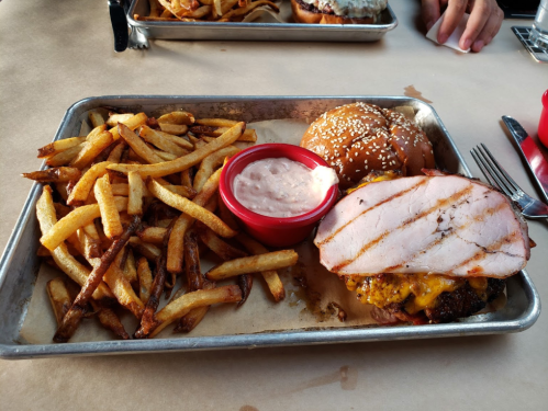 A metal tray with fries, a burger topped with ham, and a small bowl of dipping sauce.