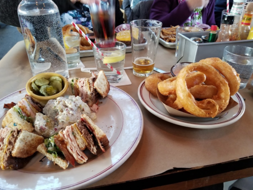 A plate with a layered sandwich, pickles, and a side of onion rings, with drinks and other food in the background.