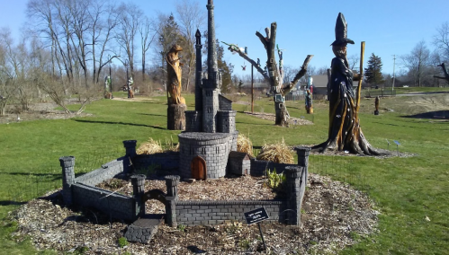 A miniature castle surrounded by a stone wall, set in a grassy area with carved wooden figures in the background.