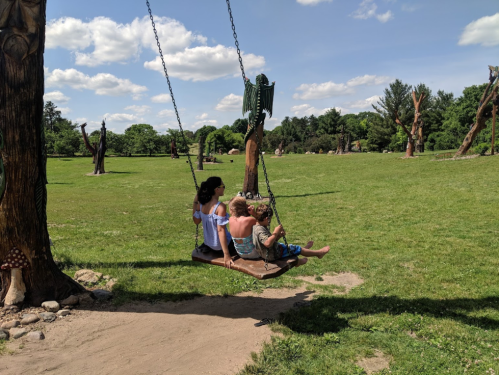 Two children and an adult swing together in a grassy park with trees and sculptures in the background under a blue sky.