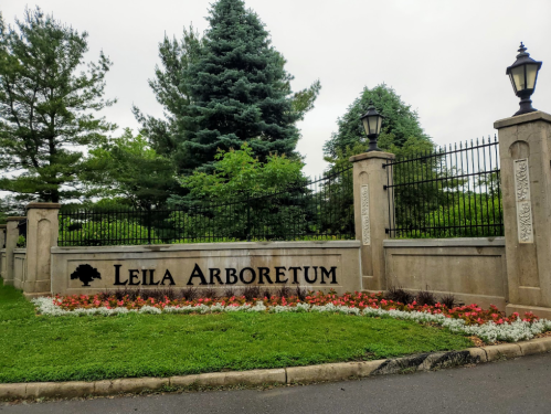 Sign for Leila Arboretum surrounded by greenery and colorful flowers, with decorative stone pillars and lamps.