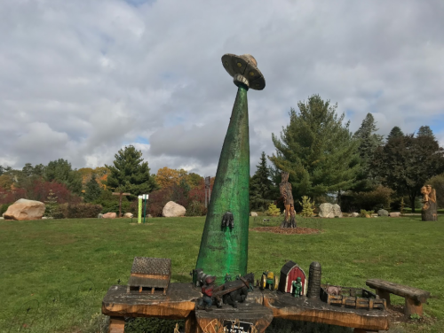 A green UFO sculpture atop a cone-shaped base, surrounded by trees and other wooden carvings in a grassy park.