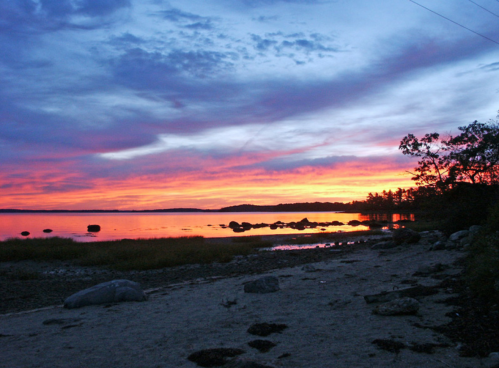 A vibrant sunset over a calm lake, with colorful clouds reflecting on the water and rocky shoreline in the foreground.