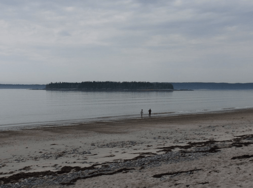 Two people walk along a sandy beach, with a calm sea and a distant island under a cloudy sky.
