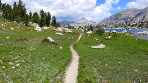 A winding dirt path through a lush green landscape, surrounded by rocks and trees, with mountains and a lake in the background.