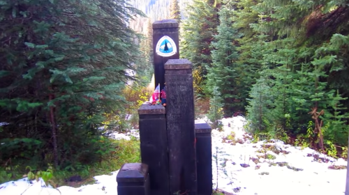A wooden monument surrounded by trees, featuring a blue emblem at the top and small decorations at its base.