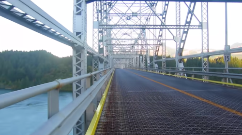 A metal bridge spans a river, with a clear sky and forested hills in the background, viewed from the bridge's surface.