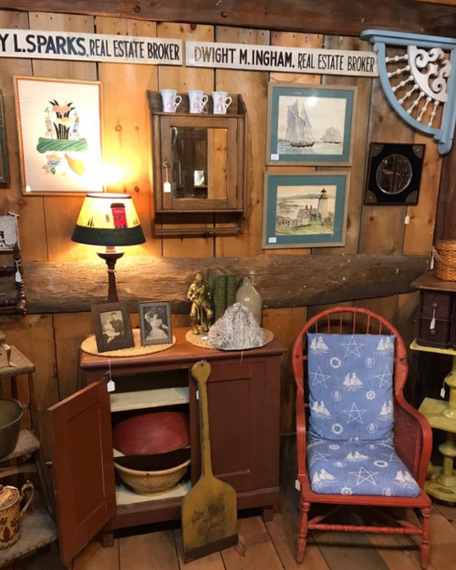 A cozy vintage shop display featuring a lamp, framed art, a chair, and various decorative items on wooden walls.