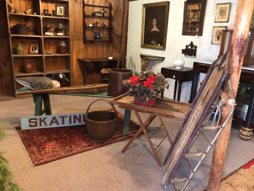 A cozy antique shop display featuring vintage furniture, decor, and a sled, with a floral centerpiece on a table.