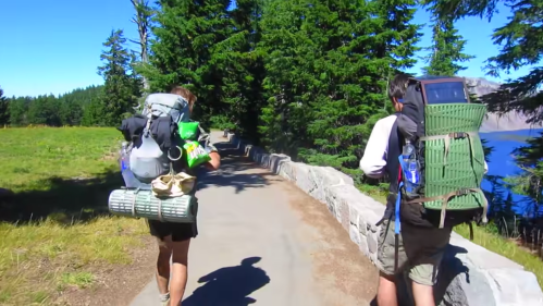Two hikers with backpacks walk along a scenic path by a lake, surrounded by tall trees and clear blue skies.