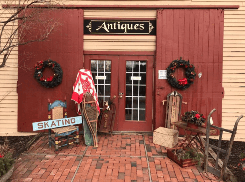 A charming antique shop entrance with festive wreaths, vintage sleds, and a sign for skating.