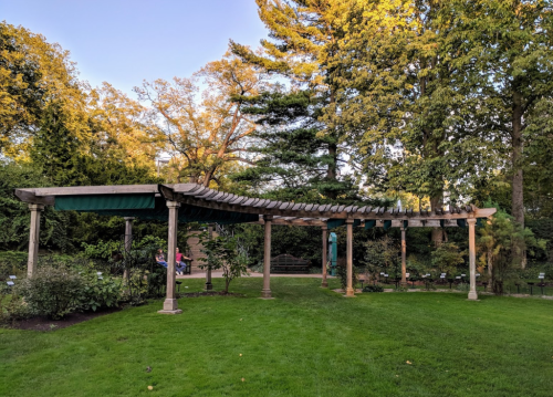 A curved wooden pergola with green fabric, surrounded by lush trees and grass in a serene garden setting.