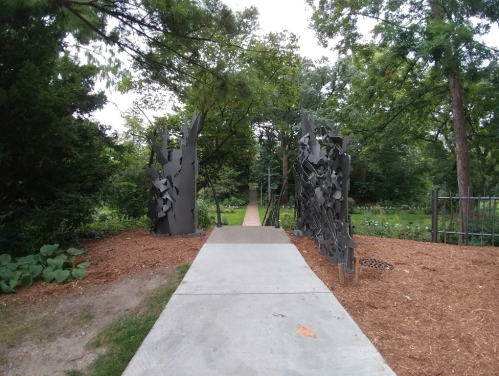 A pathway leads through abstract metal sculptures into a green garden area, surrounded by trees and mulch.