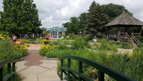 A vibrant garden with colorful flowers, pathways, and a gazebo, under a partly cloudy sky. People stroll through the area.