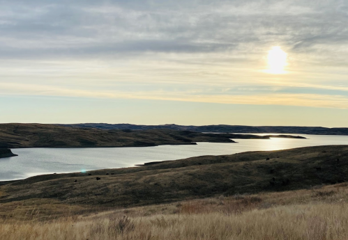A serene landscape featuring rolling hills, a calm lake, and a soft sunset sky with scattered clouds.