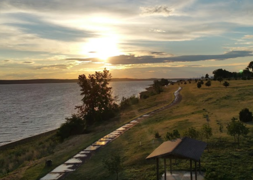 A serene lakeside view at sunset, with a path winding through green grass and trees under a colorful sky.