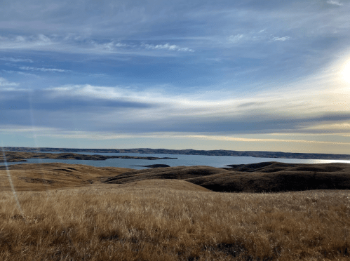 A scenic view of rolling hills and a calm lake under a blue sky with wispy clouds. Sunlight glimmers on the water.