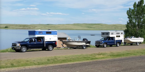 Two trucks with campers and boats are parked by a lake, surrounded by green hills and a clear blue sky.
