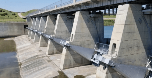 A concrete dam with large gates and supports, surrounded by water and grassy hills in the background.