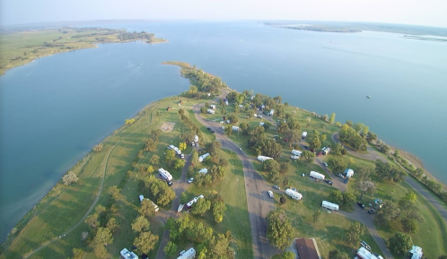 Aerial view of a lakeside campground with RVs, green trees, and a calm blue lake in the background.