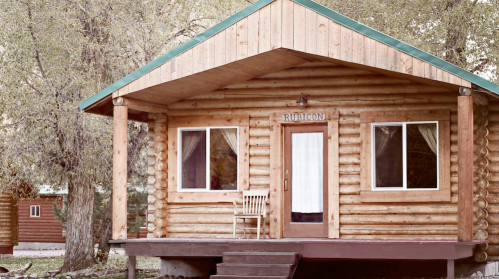 A rustic log cabin with a green roof, porch, and large windows, surrounded by trees.