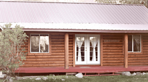 A rustic log cabin with a metal roof, featuring a front porch and large glass doors. Greenery surrounds the entrance.