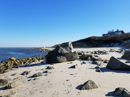 A serene beach scene with large rocks, gentle waves, and a clear blue sky, featuring a distant house on the shore.