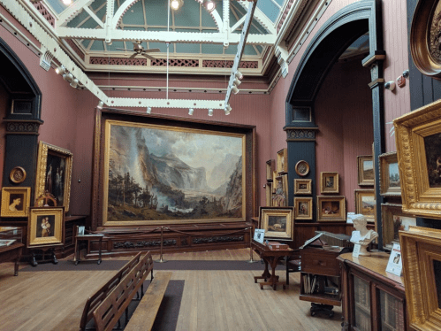 Interior of an art gallery featuring large paintings, ornate frames, and wooden benches in a historic setting.
