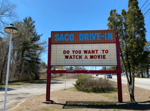 Sign at Saco Drive-In reads, "Do you want to watch a movie?" with trees and a road in the background.