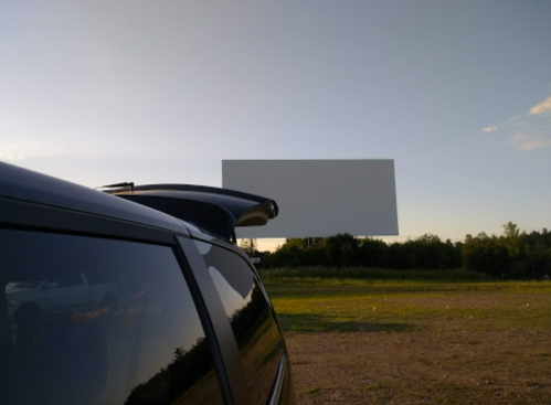 A blank movie screen stands in a field, with a car's roof visible in the foreground under a clear sky.