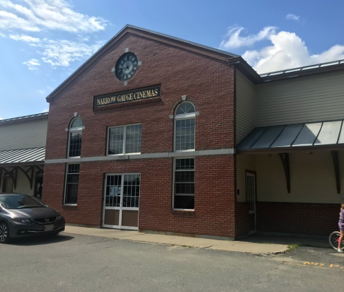Exterior of Narrow Gauge Cinemas, a brick building with large windows and a clock, under a blue sky.