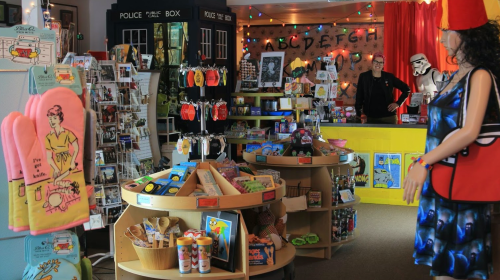 A colorful gift shop filled with quirky items, toys, and decorations, featuring a mannequin and a person at the counter.