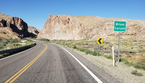 A winding road leads to a sign for Wilson Canyon, surrounded by rocky cliffs and desert landscape.