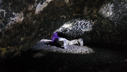 A person crawls through a dark cave, illuminated by a beam of light, showcasing rugged rock formations.