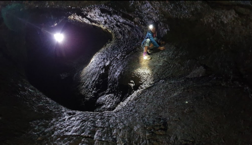 A person climbs a rocky cave illuminated by two bright lights, showcasing the cave's textured walls and winding path.