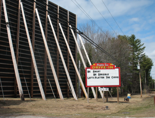 A drive-in theater sign reads "Be Smart, Be Sensible, Let's Flatten The Curve" with a backdrop of trees and a cloudy sky.