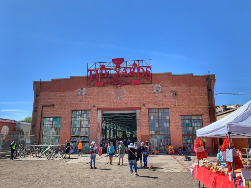 A large brick building with "The Yards" sign, bustling with people and vendors on a sunny day.