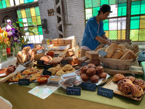 A baker arranges a variety of fresh breads and pastries on a table, surrounded by colorful stained glass windows.