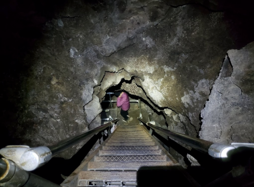 A person descends a metal staircase into a dark cave, illuminated by a flashlight, revealing rocky walls.