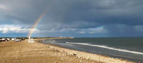 A beach scene with a rainbow arching over dark clouds and calm waves, with people and vehicles in the distance.