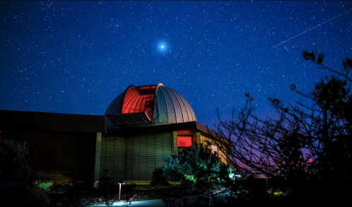 A dome-shaped observatory under a starry night sky, illuminated by red light, with plants in the foreground.