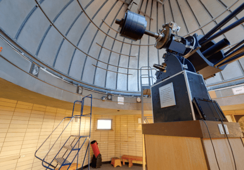 Interior of an observatory featuring a large telescope, wooden walls, and a staircase leading to the observation area.