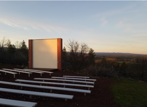 An outdoor amphitheater with white benches facing a blank screen, surrounded by trees and a sunset view in the background.