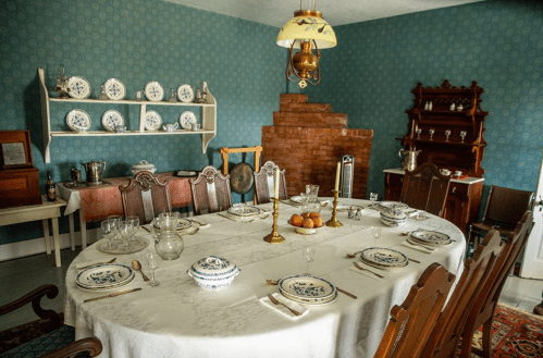A vintage dining room with a large table set for a meal, blue wallpaper, and decorative plates on a shelf.